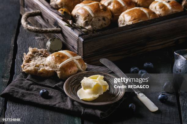 Hot cross buns in wooden tray served with butter, knife, blueberries, easter eggs, birch branch, jug of cream on textile napkin over old texture wood...