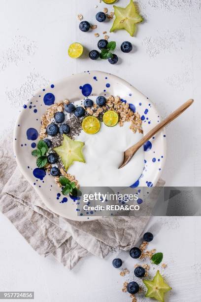 Smoothie bowl healthy breakfast. Yogurt with blueberries, granola, chia seeds, lime, wooden spoon, mint and carambola on textile napkin over white...