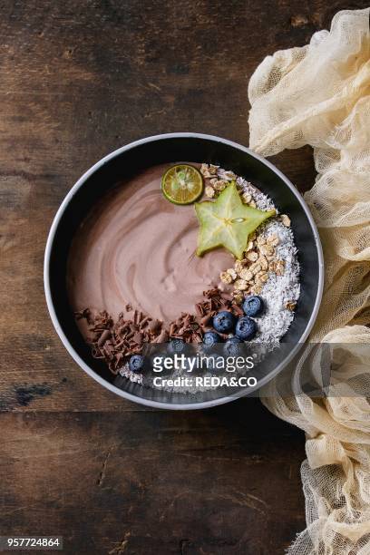 Smoothie bowl healthy breakfast. Chocolate yogurt with blueberries, granola, coconut, lime and carambola on textile gauze over dark wooden texture...