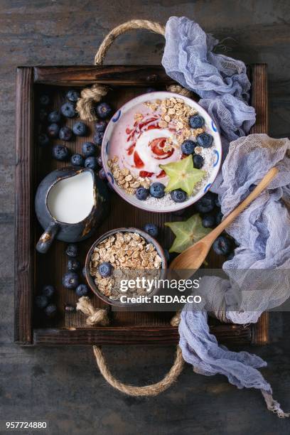 Smoothie bowl healthy breakfast. Strawberry yogurt with blueberries, granola, mint, jug of milk and carambola in wooden tray on textile gauze over...