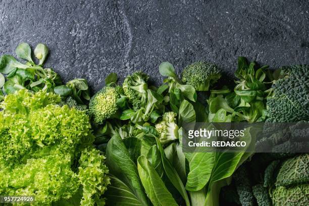 Variety of raw green vegetables salads, lettuce, bok choy, corn, broccoli, savoy cabbage as frame over black stone texture background. Top view,...