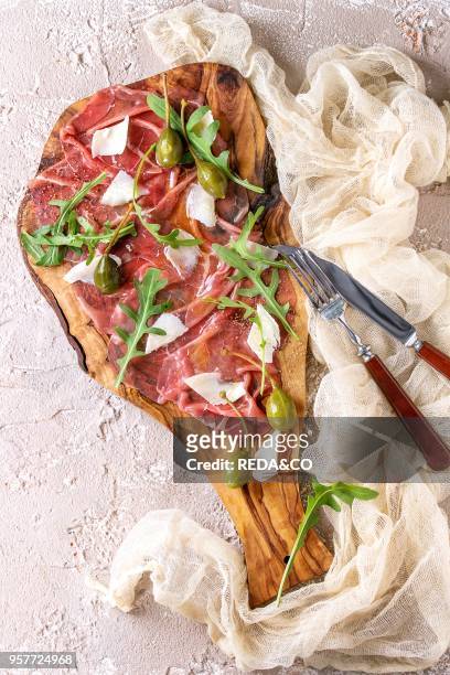 Beef carpaccio on olive wood serving board with capers, olive oil cheese and arugula, served with knife over beige concrete texture background. Top...