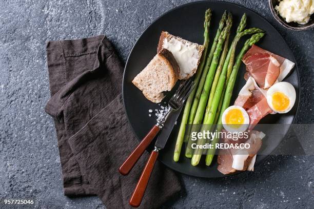 Cooked green asparagus with half boiled egg, sliced bread, butter and ham bacon served with sea salt and cutlery on black ceramic plate over dark...