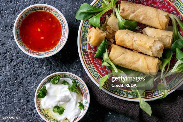 Fried spring rolls with red and white sauces, served in traditional china plate with fresh green salad over black texture background. Flat lay,...