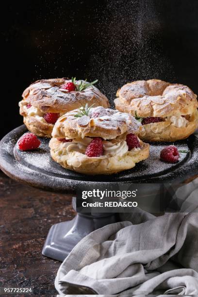 Homemade choux pastry cake Paris Brest with raspberries, almond and rosemary, served on black wooden serving board on cake stand over dark texture...