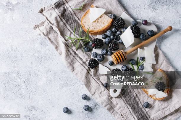 Berries blackberry and blueberry, honey on dipper, rosemary, sliced goat cheese with bread served on ceramic board with textile linen over gray...