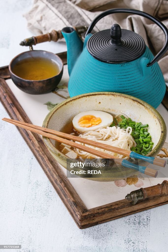 Bowl of asian style soup with noodles, egg, spring onion and tofu cheese, served with chopsticks, teapot and cup of green tea on wood tray with textile napkin, over white wooden background.