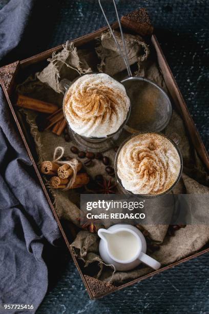 Pumpkin spicy latte with whipped cream and cinnamon in two glasses standing in wooden box with textile, jug of cream and spices other dark...