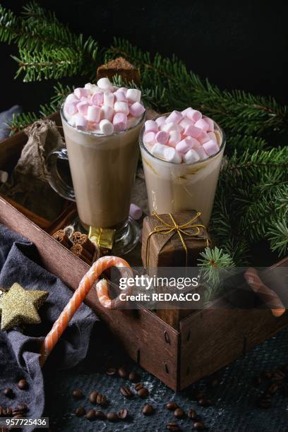 Two glasses of cafe latte with pink marshmallow standing in wooden box with Christmas decor, candies, spices, coffee beans and fir tree over dark...