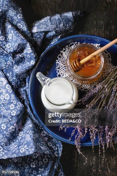 Open glass jar of liquid honey with honeycomb and honey dipper inside, jug of milk and bunch of dry lavender in blue plate over old wooden surface...