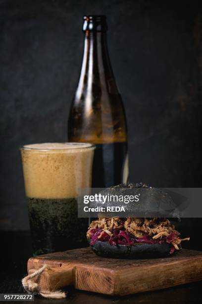 Black homemade burger with stews and red cabbage served on small wooden chopping board with glass and bottle of dark beer over wooden table with...