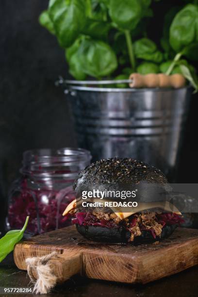 Black burger with beef stews, cheese, red cabbage and balsamic sauce served on small wooden chopping board with fresh basil over wooden table with...
