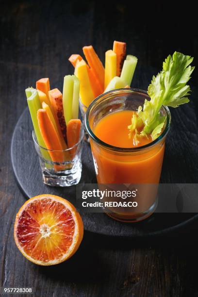 Sliced colorful raw carrots and celery as vegetarian snack, blood oranges with glass cup of fresh orange and carrot juice over black wooden table...