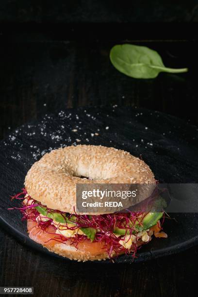 Bagel with salted salmon, spinach, beetroot sprouts, avocado and scrambled egg on wooden chopping board over old wood black background.