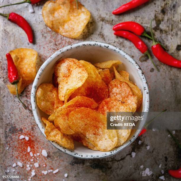 Bowl of spicy potato chips with sea salt and red hot chili peppers fresh and powder over old textural metal background. Top view. Unhealthy eating...