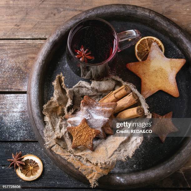 Glass mug of hot red mulled wine spices, sugar shortbread cookies star shape, anise and cinnamon powder in clay tray over dark wooden background....