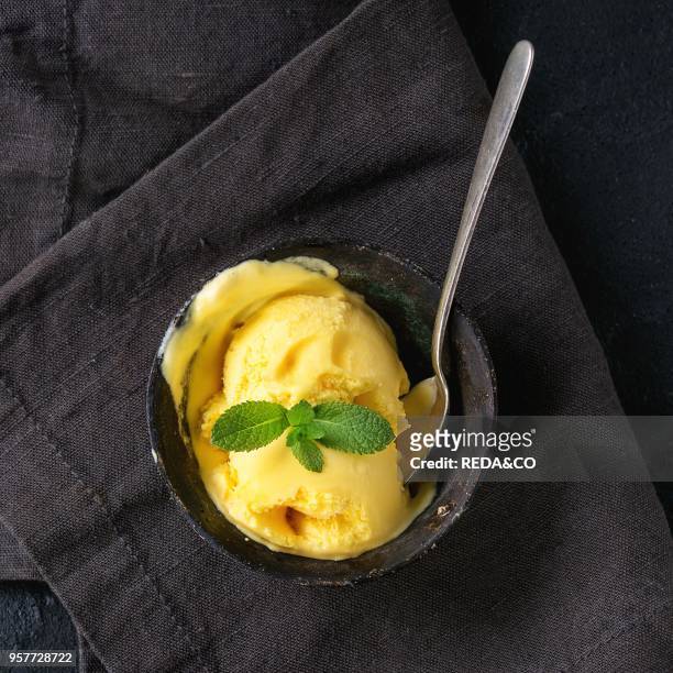 Homemade mango ice cream with fresh mint in vintage iron bowl on dark textile napkin over black textured background. Flat lay. Square image.