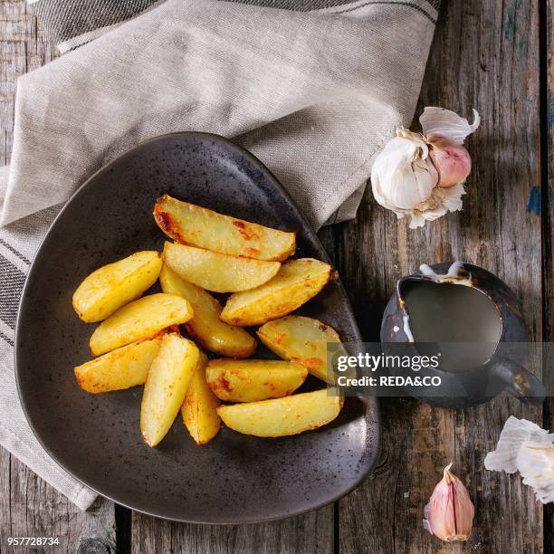 Fried country potatoes on brown ceramic plate, served with homemade aioli sauce in jug, garlic and vintage fork on gray linen napkin over wooden...