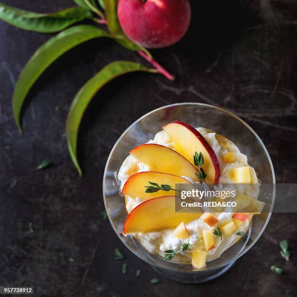 Ricotta dessert with sliced peach, thyme and honey, served in cocktail glass with whole peaches and leaves over dark textured background. Summer...