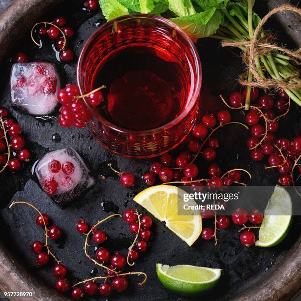 Ingredients for making lemonade Ice cubes, red currant berries, lime, lemon and mint, served with glass of juice on clay tray with black napkin over...
