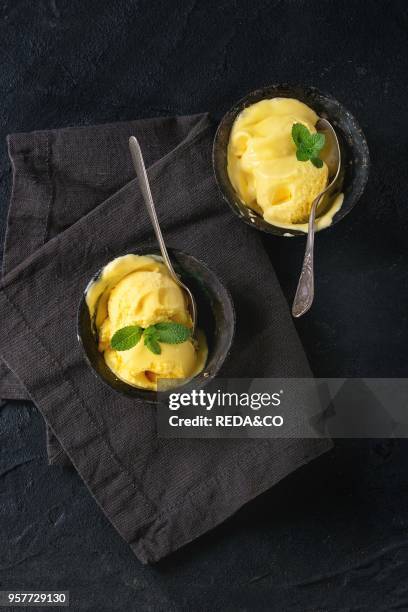 Homemade mango ice cream with fresh mint in vintage iron bowls on dark textile napkin over black textured background. Flat lay.