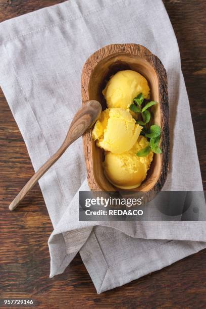 Homemade mango ice cream with fresh mint in olive wood bowl with wooden spoon, served on white textile napkin over wooden textured background. Flat...