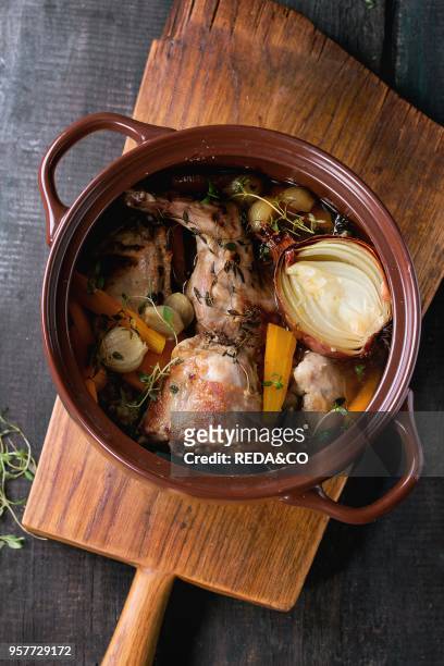 Brown ceramic pot with stewed rabbit with bouillon, vegetables and herbs, served on wooden chopping board over old wood table. Rustic style. Flat lay.