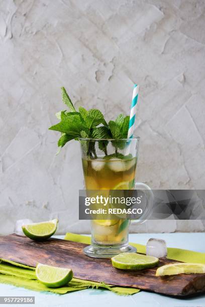 Glass of Iced green tea with lime, lemon, mint and ice cubes on wooden chopping board over light blue textured background with green textile napkin.
