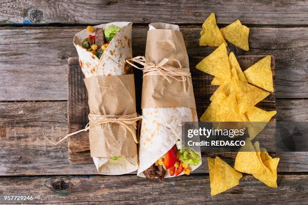 Mexican style dinner. Two papered tortillas burrito with beef and vegetables served with nachos chips over old wooden background. Flat lay.