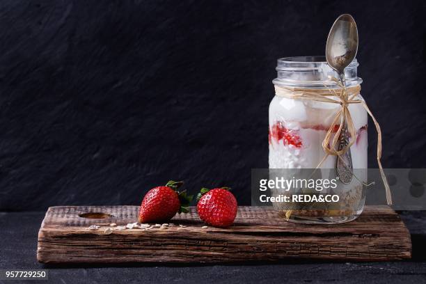 Healthy breakfast muesli, strawberries and yogurt in glass mason jar with spoon. Yoghurt served on wooden chopping board with black textured wall at...