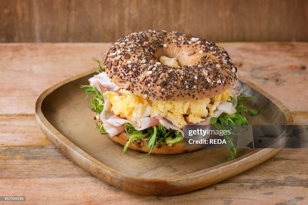 Sprinkle seeds Whole Grain bagels with scrambled eggs, pea sprout and prosciutto ham on wood plate over wooden textured background.