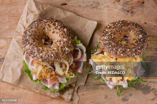 Two Whole Grain bagels with fried onion, scrambled eggs, green salad and prosciutto ham on paper over wooden textured background. Top view.