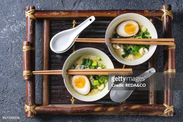 Two bowls with asian style soup with scrambled eggs, half of marinated egg, spring onion, spinach served with wood chopsticks and spoons on bamboo...