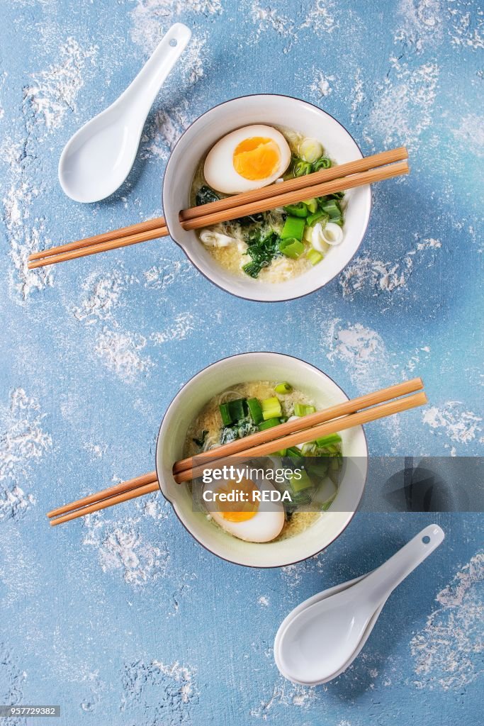 Two bowls with asian style soup with scrambled eggs, half of marinated egg, spring onion, spinach served with wood chopsticks and spoons over blue texture concrete background. Top view with space