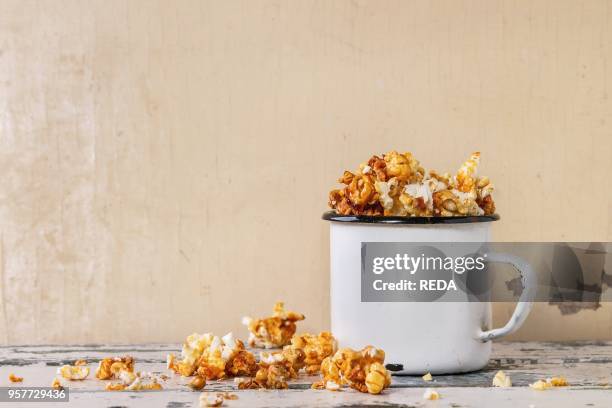 Prepared caramelized sweet popcorn served in vintage white enameled mug over old white wooden background. With space for text.