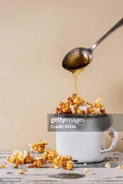 Prepared caramelized sweet popcorn served in vintage white enameled mug over old white wooden background. Caramel sause is poured from spoon. With...