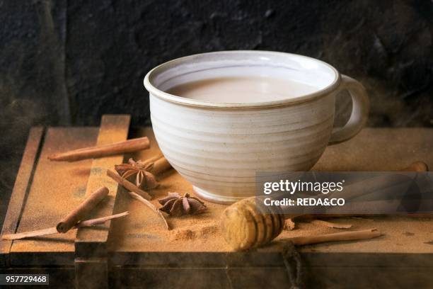 White ceramic cup of hot masala chai, served with honey on honey dipper, anise, cinnamon sticks and powder over black wooden box over black textured...