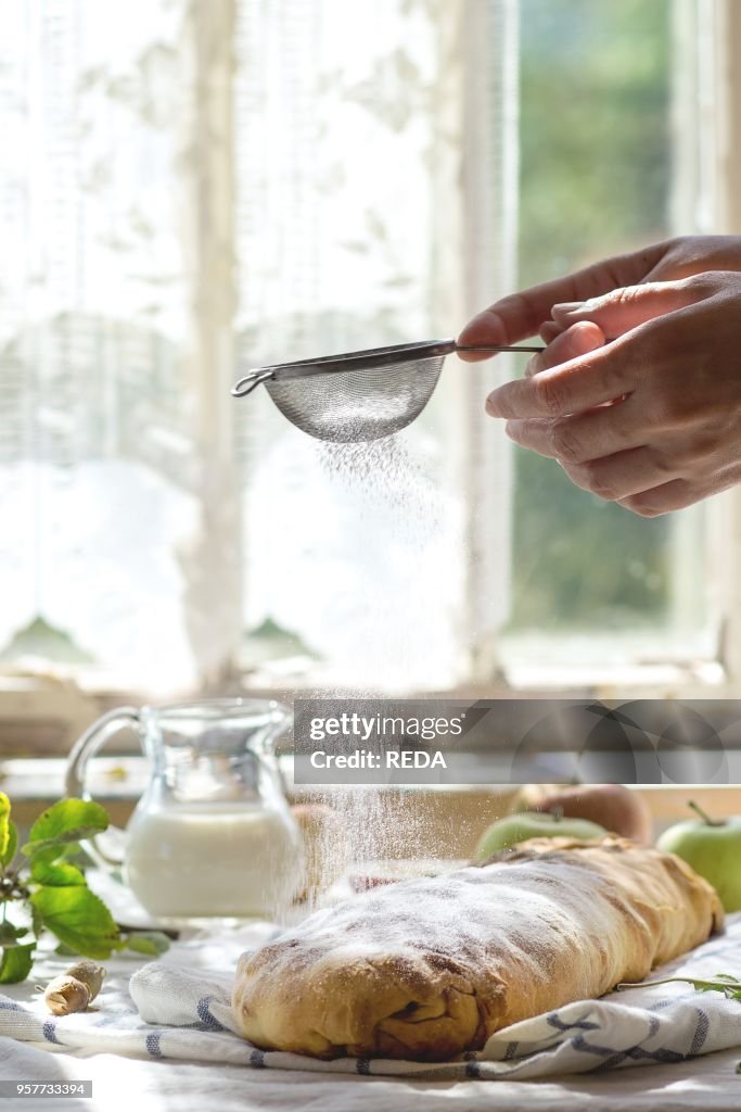 Woman's hand sprinkle with powdered sugar fresh baked homemade apple strudel over towel on kitchen table with jug of milk and apples. With window as background. Rustic style. Natural day light.
