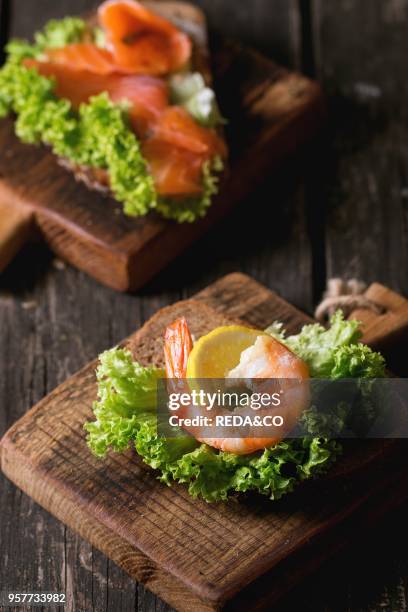 Sandwiches with whole wheat bread, fresh salad, shrimp and salted salmon on little wooden cutting board over old wooden table. Rustic style.