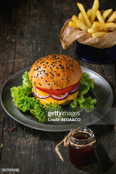 Fresh homemade hamburger with black sesame seeds in old metal plate with fried potatoes, served with ketchup sauce in glass jar over old wooden table...