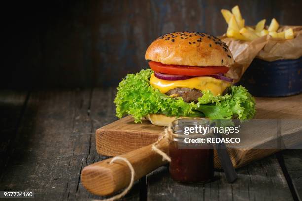 Fresh homemade burger with black sesame seeds on wooden cutting board with fried potatoes, served with ketchup sauce in glass jar over old wooden...