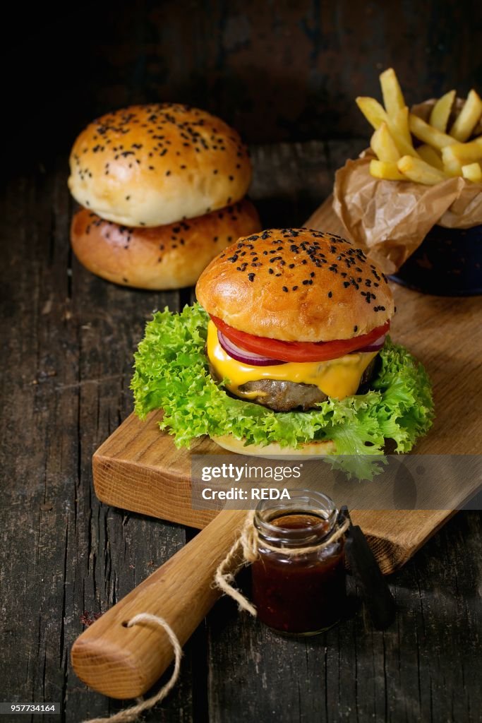 Fresh homemade burger with black sesame seeds on wooden cutting board with fried potatoes, served with ketchup sauce in glass jar over old wooden table with dark background. Dark rustic style.