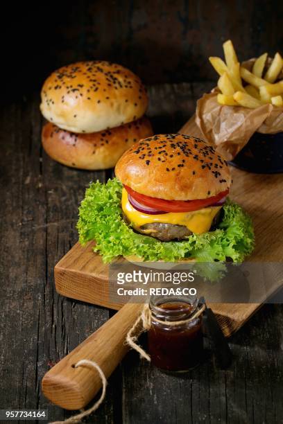 Fresh homemade burger with black sesame seeds on wooden cutting board with fried potatoes, served with ketchup sauce in glass jar over old wooden...