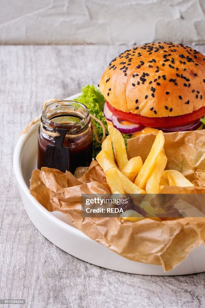 Fresh homemade burger with black sesame seeds in white plate with fried potatoes, served with ketchup sauce in glass jar over gray wooden table.