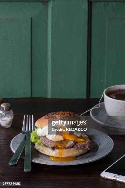 Cafe Breakfast with homemade sandwich with baked meat and soft-boiled egg, cup of hot pocket tea and white smartphone over dark wooden table with...