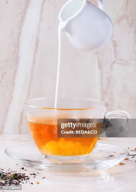 Milk pouring from white jug to glass cup of hot tea on saucer with dry green and black tea leaves over white marble backgtound.