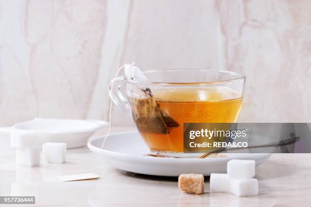 Glass cup of hot tea on saucer with sugar cubes, spoon and tea bag over white marble backgtound. Top view.