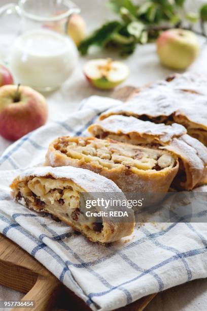 Sliced fresh baked homemade apple strudel over towel on kitchen table with jug of milk and apples. Rustic style. Natural day light.