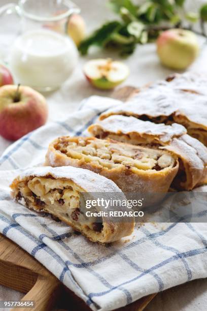 Sliced fresh baked homemade apple strudel over towel on kitchen table with jug of milk and apples. Rustic style. Natural day light.