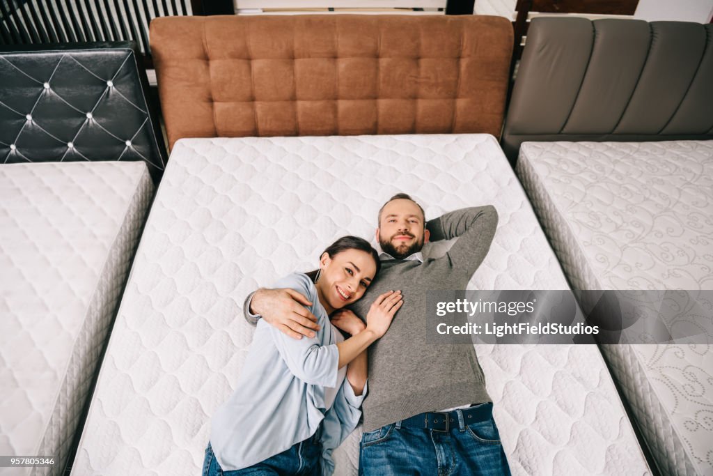 Overhead view of smiling couple lying on bed in furniture store with arranged mattresses